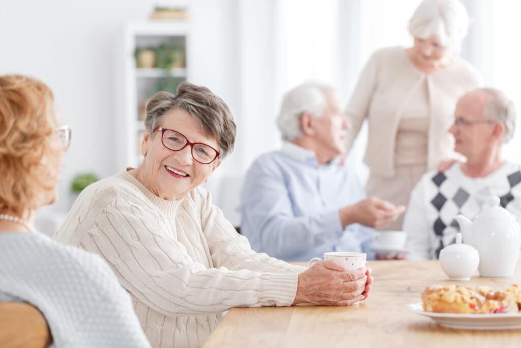 A group of older adults sit and stand around a wooden table, chatting and smiling. One woman in the foreground, wearing glasses and a cream sweater, holds a mug. Pastries and a teapot are on the table.