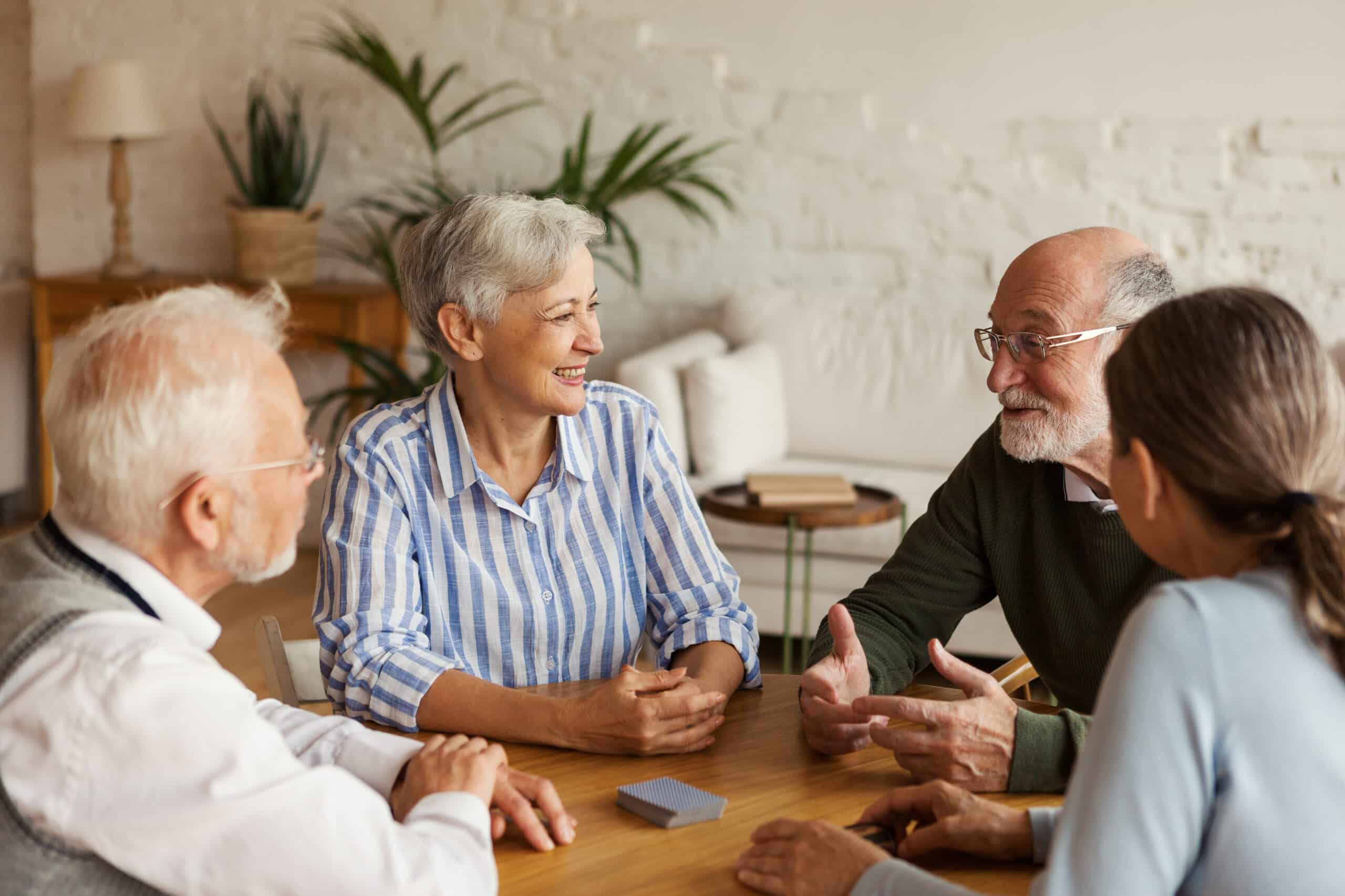 Four older adults sit around a table, smiling and talking with each other in a bright, cozy room with plants and books in the background. A deck of cards is on the table.