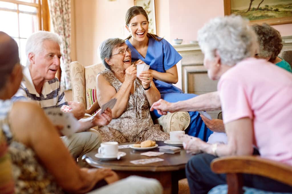 A group of elderly people sit around a table playing cards and laughing with a young female caregiver in blue scrubs. Teacups, plates, and cards are on the table in a cozy, sunlit room.