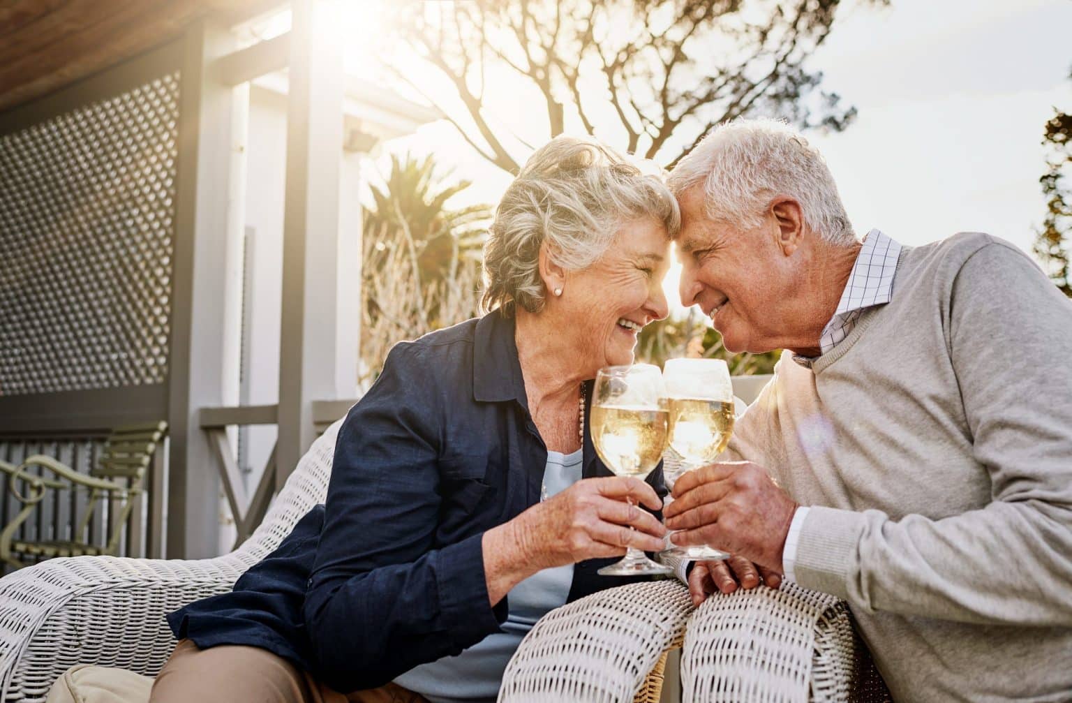 An elderly couple sits closely together on outdoor chairs, smiling and touching foreheads as they clink glasses of champagne, enjoying a sunny day on a patio.